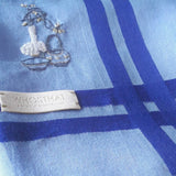 Close-up of sky blue cotton hanky, royal blue stripes border, and the delicate hand embroidery of Chinese tea setting next to vase with flowers. 