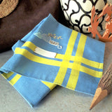 Blue cotton handkerchief with yellow stripes as border, has a delicate hand embroidery of a vase with zen style flower arrangement surrounded by digital clock, and a tea pot.