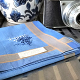 Ocean blue cotton pocket square is hand embroidered a flying plane and ancient huts in the greens