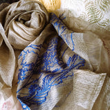 Close up of sheer and glimmering olive color silk wool scarf has a big blue line art landscape pattern near the hem 