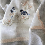 Close-up of a light grey super fine cashmere scarf decorated with velveteen beige stripes, hand embroidered two blue birds stand on branches of white flowerings