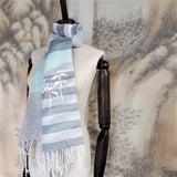 A sheer cotton linen scarf in blue and navy color stripes has a white straw tape embroidered pine trees pattern and  white fringe at the hem.