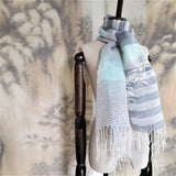 A sheer cotton linen scarf in white, navy and icy blue color stripes has three white straw tape embroidered pine trees pattern and hand twisted fringe at the hem.