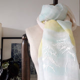Close up on a sheer cotton linen scarf in white, yellow and icy blue color stripes has three white straw tape embroidered pine trees pattern